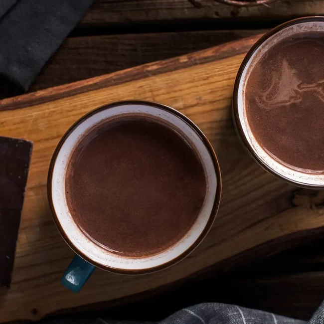 Topla čokolada u dve šolje na drvenoj dasci i komad čokolade pored šolja.