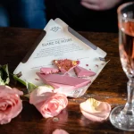 Aguara ruby čokolada sa jagodom, malinom i kajsijom.Na stolu se nalazi izlomljena čokolada na pakovanju, roze vino i latice ruže.