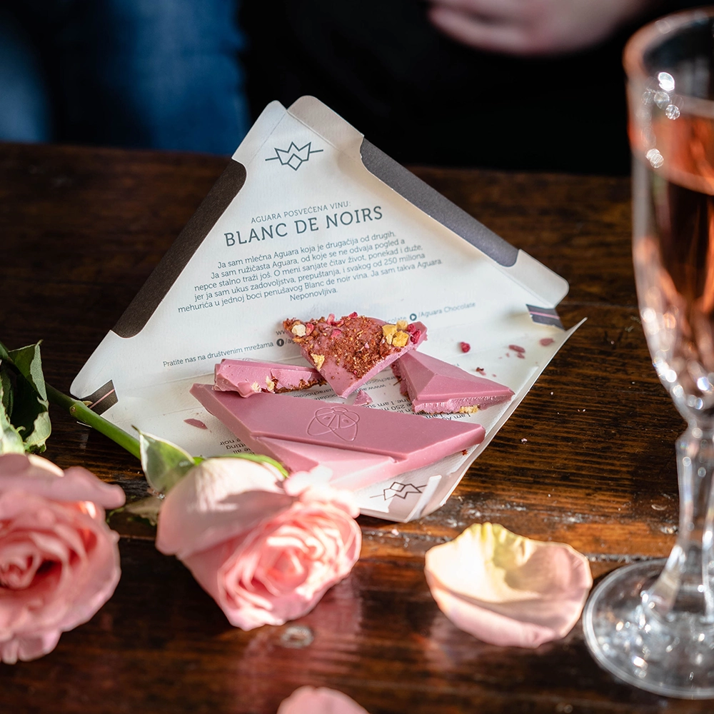 Aguara ruby čokolada sa jagodom, malinom i kajsijom.Na stolu se nalazi izlomljena čokolada na pakovanju, roze vino i latice ruže.